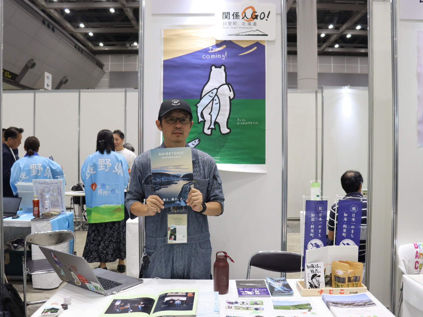 初海さんが手にしているのが『SHIRETOKO! SUSTAINABLE 海と、森と、人。』で、写真家・石川直樹氏が編集長。後ろのポスターに映っているのが地域のシンボルキャラクター「知床トコさん」